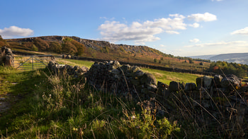 This landscape photograph captures Curbar Edge, a prominent gritstone escarpment located in Derbyshire, United Kingdom, within the Peak District National Park. Taken in the afternoon during autumn, the image showcases the natural beauty of the rural surroundings, with warm sunlight illuminating the rugged terrain and seasonal colors on the hillside. In the foreground, a traditional dry stone wall runs through grassy fields, characterizing the region’s agricultural heritage. Mature trees with changing leaves are scattered across the landscape, and the expansive view of Curbar Edge forms a dramatic backdrop, highlighting the distinct features of this nature-rich area in the heart of the Peak District.